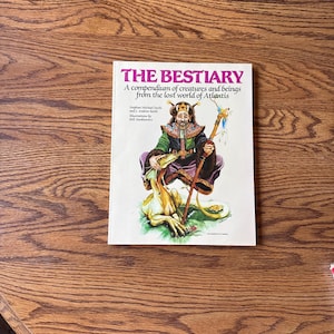 Op de afbeelding: Een boek getiteld "THE BESTIARY" met een kleurrijke illustratie van een geklede figuur die een staf vasthoudt, met een wezen aan hun voeten. De boekomslag bevat tekst over wezens en wezens uit Atlantis.