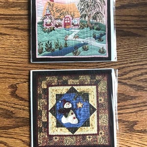 May include: Two quilted art cards. The top card features a cottage scene with a red door, a stream, and a tree. The bottom card has a snowman in a blue diamond, surrounded by a patterned border. Both cards are in protective packaging.