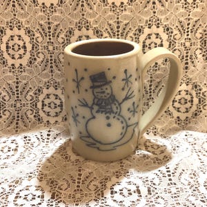 Puede incluir: Una taza de cerámica color crema con un diseño de muñeco de nieve azul. La taza tiene un interior marrón y un asa curvada. El muñeco de nieve lleva un sombrero de copa y está rodeado de copos de nieve. La taza está sobre un mantel de encaje blanco.