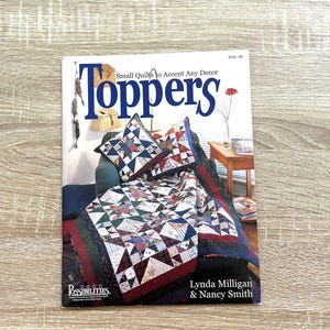 Pode incluir: Um livro de instruções de capa mole intitulado Toppers: Small Quilts to Accent Any Decor por Lynda Milligan e Nancy Smith. A capa apresenta uma fotografia de uma colcha de retalhos e almofadas a condizer com padrões de estrelas.