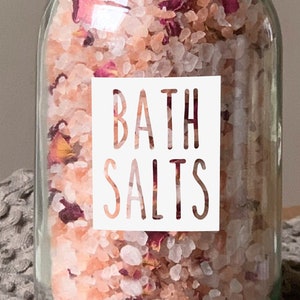May include: A clear glass jar with a black lid filled with pink bath salts and dried rose petals. A white label with the words "BATH SALTS" is affixed to the front of the jar. The jar sits on a gray, textured surface.
