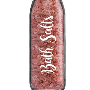 May include: A clear glass bottle filled with pink bath salts. The bottle has a silver lid and a white label that says "Bath Salts".