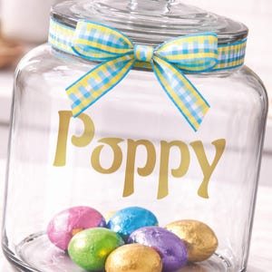 Peut inclure: Un bocal en verre transparent avec un couvercle et un ruban vichy bleu et jaune noué autour du haut. Le mot "Poppy" est en lettres dorées sur le devant. À l'intérieur du bocal, plusieurs œufs en chocolat emballés dans du papier d'aluminium de différentes couleurs.