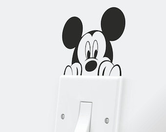 Op de afbeelding: Een witte lichtschakelaar met een zwarte Mickey Mouse-sticker die over de bovenkant uitsteekt. De sticker toont Mickey's oren, ogen en neus.
