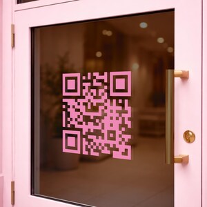Peut inclure: Une porte rose avec une fenêtre en verre ornée d'un grand autocollant de code QR rose. Le cadre de la porte et la quincaillerie sont également roses, avec des poignées et une serrure dorées. L'arrière-plan est flou.
