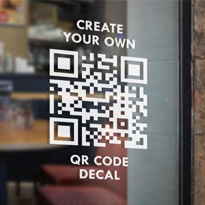 May include: A white QR code decal on a glass surface. The decal features a large, complex QR code design, with the words "CREATE YOUR OWN" above and "QR CODE DECAL" below the code in a clean, sans-serif font. The background is blurred.