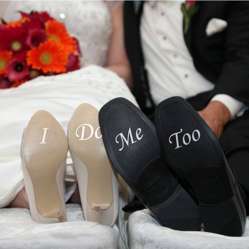 May include: A pair of white high-heeled shoes with the words "I Do" written on the soles, and a pair of black dress shoes with the words "Me Too" written on the soles.