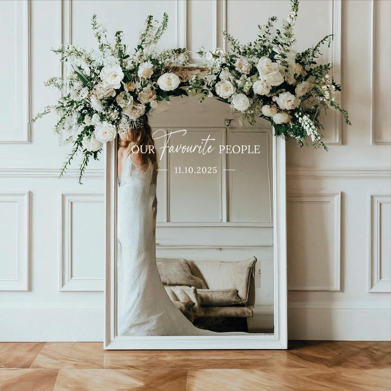 May include: A white framed mirror with the text "OUR Favourite PEOPLE 11.10.2025". The mirror is decorated with white flowers and greenery. The reflection shows a bride in a white dress and a beige sofa.