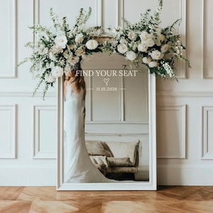 Może przedstawiać: Duże, prostokątne lustro z białą ramą, ozdobione kompozycją kwiatową z białych róż i zieleni. Lustro wyświetla tekst "FIND YOUR SEAT" i datę. Obraz sugeruje scenerię ślubną lub wydarzenie.