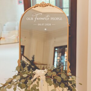 May include: A gold-framed, arched mirror with the text "OUR Favourite PEOPLE" and the date "08.07.2026" in white script. The mirror is decorated with greenery and a white bow at the base, suitable for a wedding or special event.