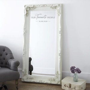 May include: A large, ornate white framed mirror with the text "OUR Favourite PEOPLE - 10.08.2026". The mirror is leaning against a white wall. A gray armchair and a small suitcase with flowers are in the foreground.