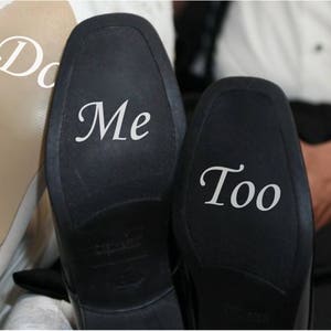May include: A pair of black leather shoes with white text on the soles. One shoe says "Me" and the other says "Too".