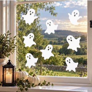 May include: A set of seven white ghost window decals featuring simple cut-out eyes and mouths. Each ghost has a rounded head and a wavy, flowing bottom edge. These adhesive decorations are arranged in a scattered pattern across the glass pane.