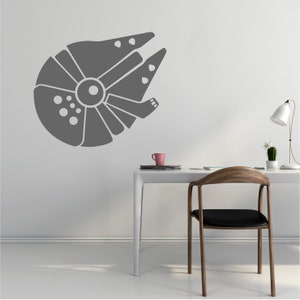 May include: A grey wall decal of the Millennium Falcon spaceship from Star Wars. The decal is on a white wall above a white desk with a wooden chair. A pink mug and a potted plant are on the desk.