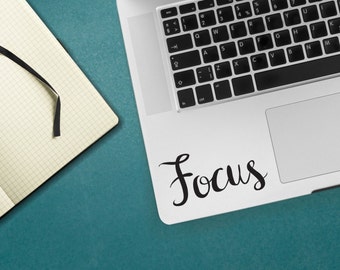 May include: A silver laptop computer with a black keyboard and a white trackpad. The trackpad has the word "Focus" written on it in black cursive lettering.