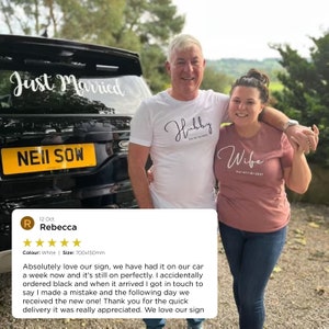 May include: A couple poses in front of a black car with a "Just Married" sign on the back window. The man is wearing a white t-shirt with the word "Hubby" written in cursive. The woman is wearing a pink t-shirt with the word "Wife" written in cursive. The car's license plate reads "NEII SOW".