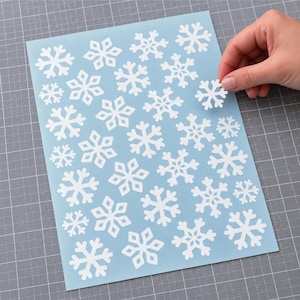 May include: A light blue sheet featuring a pattern of multiple white snowflake stickers of varying sizes. One individual white snowflake sticker is being peeled off the sheet, revealing the adhesive side against the light blue background.