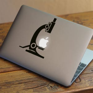 May include: A black microscope sticker on a silver laptop. The microscope is positioned over the Apple logo on the laptop.