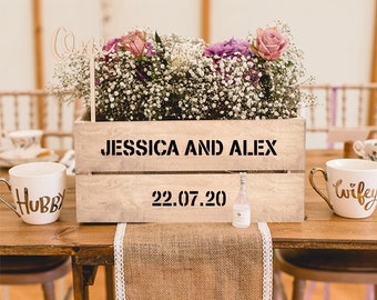 May include: A wooden crate with the words "Jessica and Alex" and the date "22.07.20" engraved on it. The crate is filled with white and pink flowers and a small glass bottle. The crate is sitting on a wooden table with a burlap runner.