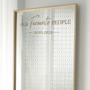 May include: A rectangular mirror with a gold frame. The mirror has the words "OUR Favourite PEOPLE" and the date "30.05.2026" in a decorative font. The background features a white geometric pattern.