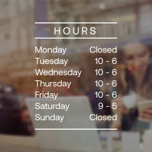 May include: White text on a glass window displays business hours. The word "HOURS" is at the top. Below, the days of the week are listed with corresponding opening and closing times. The background is blurred.