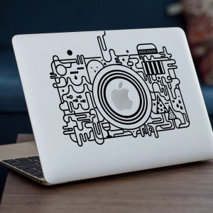 May include: A white laptop computer with a black and white abstract design sticker on the lid. The design features a stylized camera lens with an Apple logo inside.