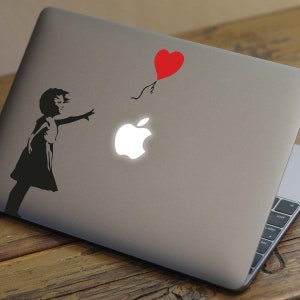 May include: A black and white silhouette of a girl reaching for a red heart-shaped balloon that is floating away. The girl is on a silver laptop with a white Apple logo.