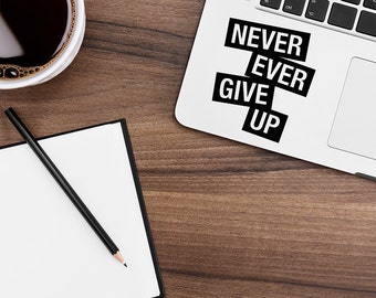 May include: A laptop with a sticker saying "NEVER EVER GIVE UP" in black text on a white background. A cup of coffee, a notepad, and a pencil are on a wooden surface.