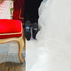 May include: A pair of black leather shoes with the words "Help" and "Me!" written on them. The shoes are on a white surface with a red chair and a white wedding dress in the background.