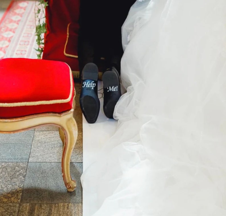 May include: A pair of black leather shoes with the words "Help" and "Me!" written on them. The shoes are on a white surface with a red chair and a white wedding dress in the background.