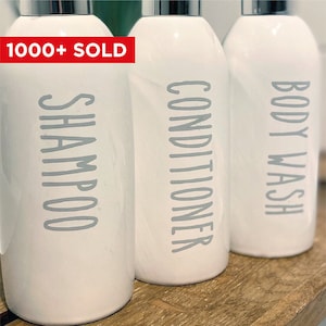 Puede incluir: Tres botellas de cerámica blanca con bomba y letras grises. Las botellas están etiquetadas como "Champú", "Acondicionador" y "Gel de baño".