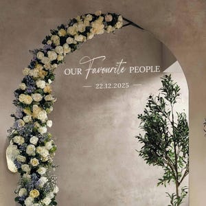 May include: An arched mirror with the text "Our Favourite People" and the date 22.12.2025. The arch is decorated with white and yellow flowers and greenery. A tree with green leaves is on the right side of the mirror.