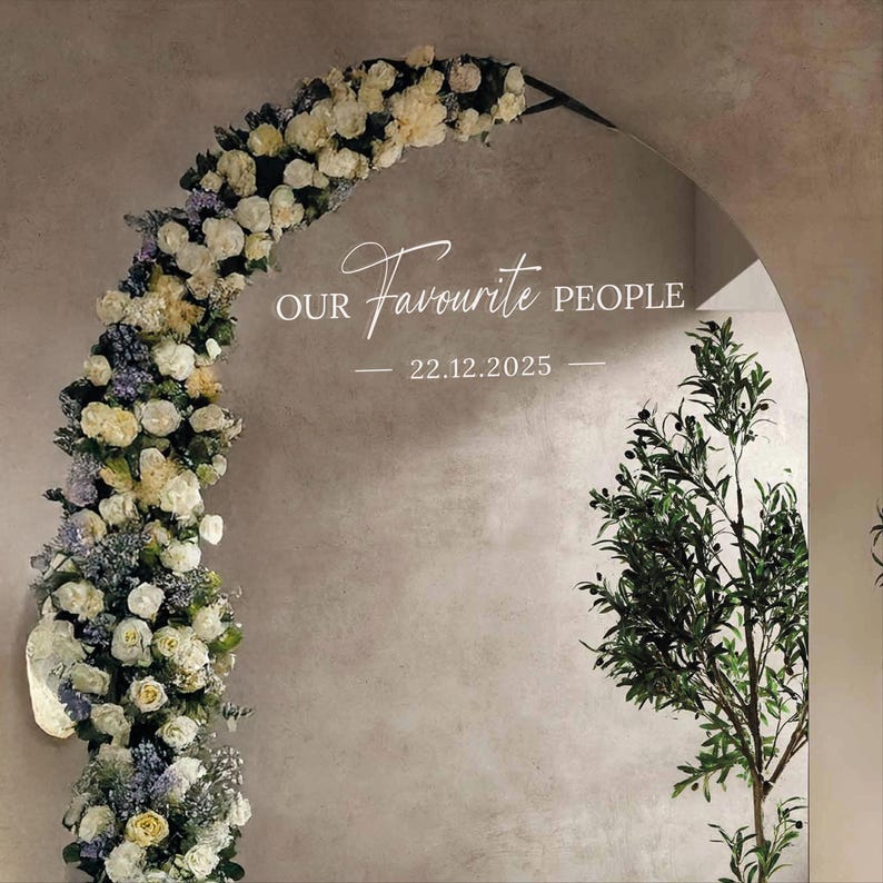 May include: An arched mirror with the text "Our Favourite People" and the date 22.12.2025. The arch is decorated with white and yellow flowers and greenery. A tree with green leaves is on the right side of the mirror.
