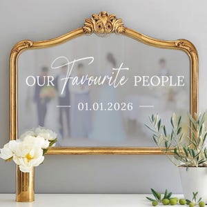 May include: A gold-framed mirror with ornate detailing at the top. The mirror's surface reflects a wedding scene and features the text "OUR Favourite PEOPLE 01.01.2026". A gold vase with white flowers and a potted olive plant are in the foreground.
