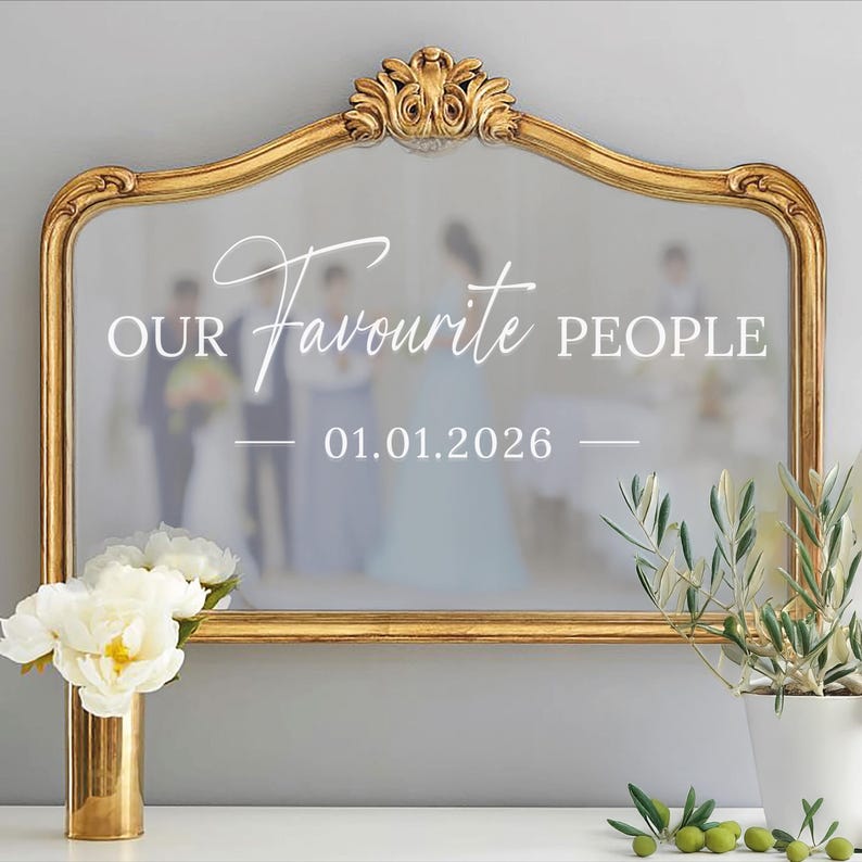May include: A gold-framed mirror with ornate detailing at the top. The mirror's surface reflects a wedding scene and features the text "OUR Favourite PEOPLE 01.01.2026". A gold vase with white flowers and a potted olive plant are in the foreground.