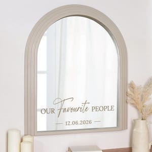 May include: A beige arched mirror with the text "OUR Favourite PEOPLE" and the date "12.06.2026". The mirror is mounted on a white wall. A vase with dried flowers and candles are visible.