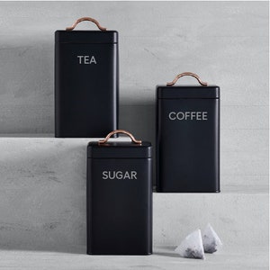 May include: Three black metal canisters with rose gold lids and handles. The canisters are labeled "TEA", "COFFEE", and "SUGAR".