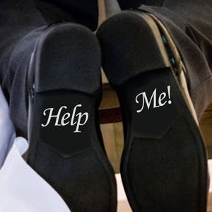 May include: A pair of black dress shoes with white text on the soles. One shoe says "Help" and the other says "Me!"
