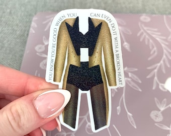 May include: A gold and black sticker with a figure wearing a gold jacket and black pants. The sticker has the text "You know you're good when you can even do it with a broken heart."