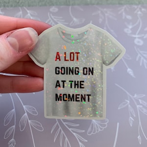 May include: A white t-shirt sticker with a glittery finish. The text on the sticker reads "A LOT GOING ON AT THE MOMENT".