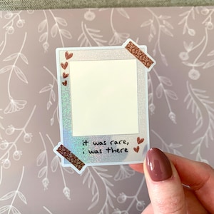 以下が含まれることがあります： 白い枠とキラキラした背景を持つホログラフィックステッカー。ステッカーは、中央に空白のある写真フレームの形をしています。ステッカーには、「it was rare, i was there」というテキストが下に書かれています。ステッカーには、茶色のハートと茶色のテープのアクセントがあります。