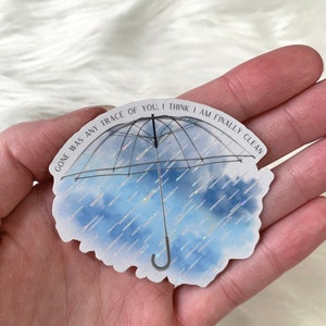 May include: A white sticker with a black outline of an umbrella with a blue and white watercolour background. The text on the sticker reads "Gone was any trace of you. I think I am finally clean."