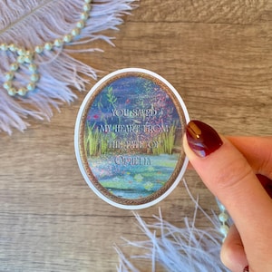 May include: An oval sticker with the text "YOU SAVED MY HEART FROM THE FATE OF OPHELIA" in white and gold. The sticker features a watercolour design of a pond with plants and flowers. The sticker is held by a hand with red nail polish.