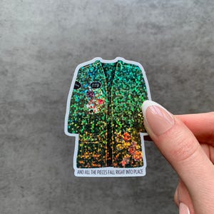 May include: A holographic sticker in the shape of a coat, featuring a green and gold glitter design. The sticker has floral accents and the text "AND ALL THE PIECES FALL RIGHT INTO PLACE". The word "rep" is also visible.
