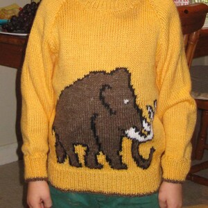 May include: A yellow knitted sweater featuring a brown woolly mammoth design. The sweater has a round neck and long sleeves with brown trim. The child is also wearing a red collared shirt and green pants.