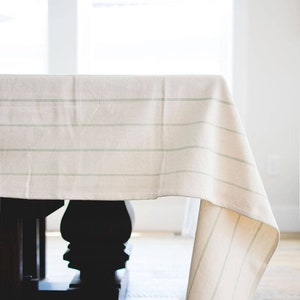 May include: A white tablecloth with thin green stripes hangs over a dark wood table. The tablecloth is draped over the table and hangs down to the floor.