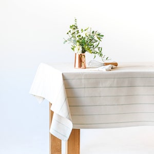 May include: A white tablecloth with thin, light green stripes, draped over a wooden table. A copper vase with a small bouquet of white flowers and greenery sits on the table, along with a whisk and a wooden utensil.