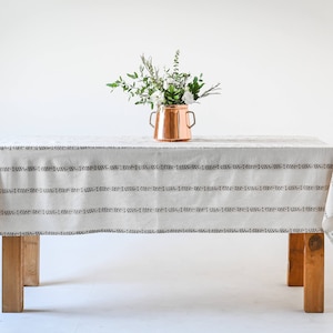 May include: A white tablecloth with a black stripe pattern, draped over a wooden table. A copper vase with a bouquet of white flowers sits on the table.