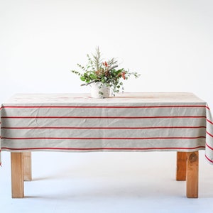May include: A rectangular wooden table with a beige tablecloth featuring red horizontal stripes. A small white vase with a floral arrangement sits on the table. The table has four wooden legs.