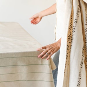 May include: A person is standing near a table with a white tablecloth with green stripes. The person is wearing a white shirt and a white shawl with a brown leaf pattern.